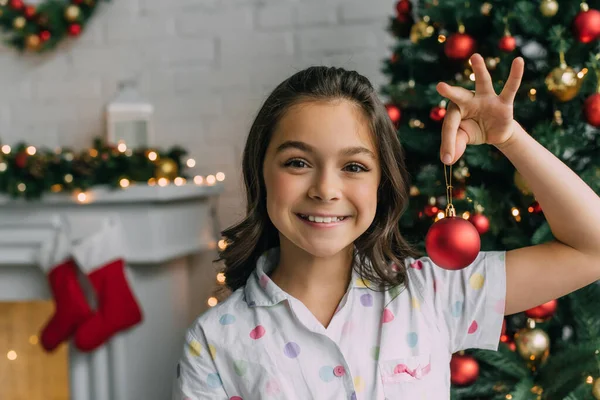 Kind im Pyjama hält Ball neben verschwommenem Weihnachtsbaum zu Hause — Stockfoto