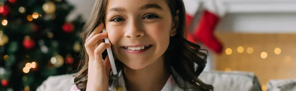 Sonriente niño mirando a la cámara mientras habla en el teléfono inteligente durante la Navidad en casa, pancarta - foto de stock