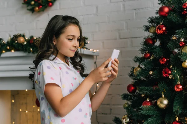 Chica preadolescente en pijama usando teléfono inteligente cerca del árbol de Navidad decorado en casa - foto de stock