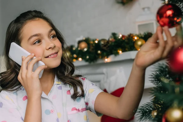 Chica preadolescente en pijama de puntos hablando en el teléfono inteligente cerca del árbol de Navidad decorado en casa - foto de stock
