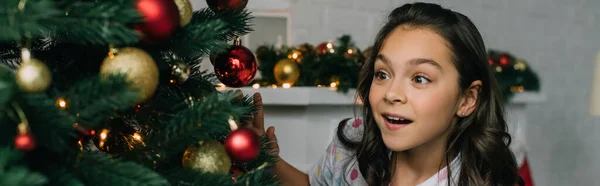 Fille excitée regardant l'arbre de Noël avec des boules à la maison, bannière — Photo de stock
