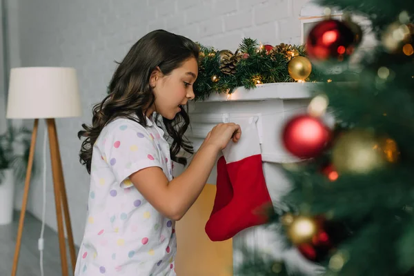 Vista laterale del bambino scioccato in pigiama guardando la calza di Natale vicino al camino a casa — Foto stock