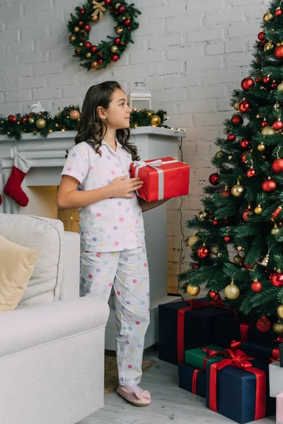 Preteen child in pyjama holding gift and looking at Christmas tree at home — Stockfoto
