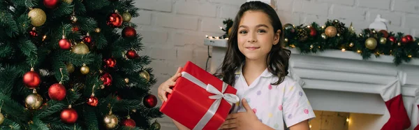 Positive girl in pajama holding present near Christmas tree in living room, banner — Stock Photo