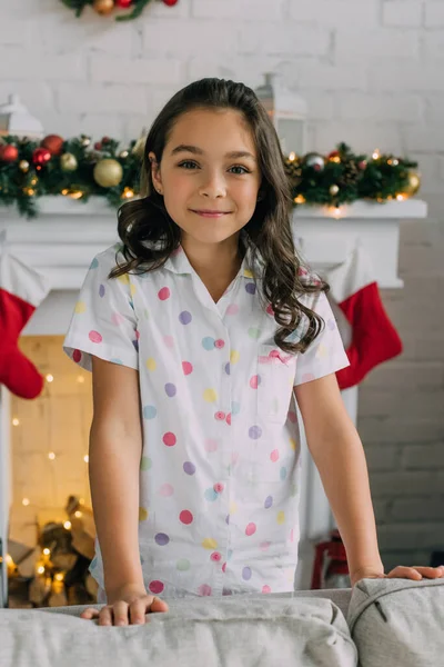 Fille heureuse en pyjama pointillé regardant la caméra près du canapé et cheminée décorée pendant les vacances d'hiver — Photo de stock