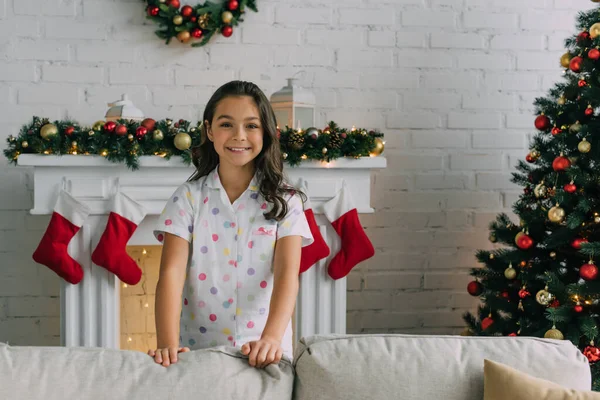 Positive kid in pajama looking at camera near Christmas decor in living room — Stock Photo