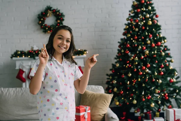 Fille positive en pyjama pointant avec les doigts près de l'arbre de Noël flou dans le salon — Photo de stock