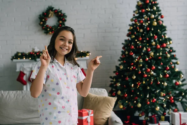 Menina positiva no pijama apontando com os dedos perto da árvore de Natal borrada na sala de estar — Fotografia de Stock