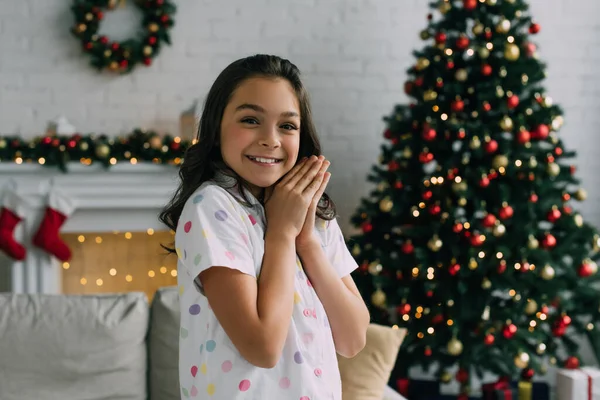 Glückliches Mädchen im Pyjama blickt in die Kamera und macht betende Hände während der Weihnachtsfeier zu Hause — Stockfoto