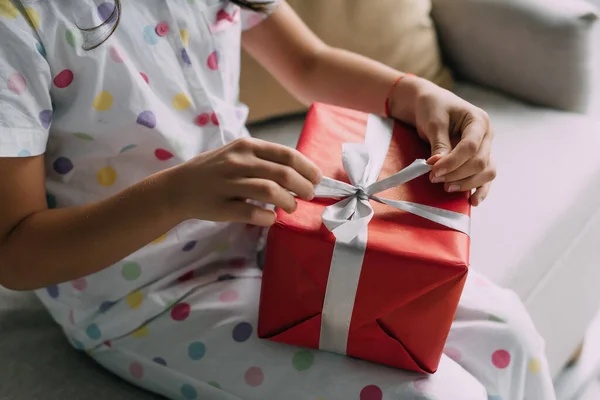 Обрезанный вид ребенка в пижаме держа рождественский подарок с луком на диване — стоковое фото