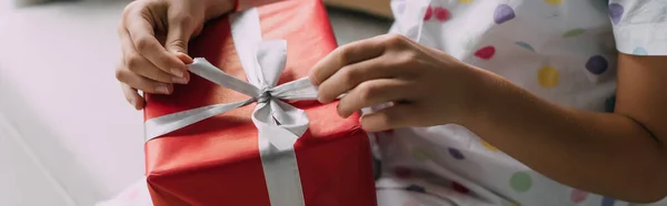 Ausgeschnittene Ansicht eines Kindes im gepunkteten Pyjama mit Weihnachtsgeschenk mit Schleife auf Couch, Banner — Stockfoto