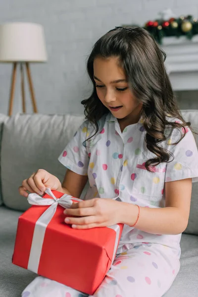 Kid in dotted pajama adjusting bow on Christmas gift at home — Stock Photo