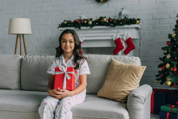 Fille souriante en pyjama pointillé tenant cadeau tout en étant assis sur le canapé pendant la célébration de Noël — Photo de stock