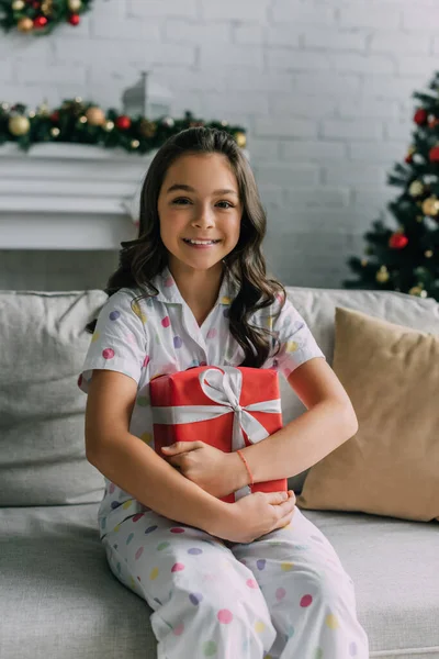 Enfant heureux en pyjama pointillé tenant cadeau du Nouvel An et assis sur le canapé — Photo de stock