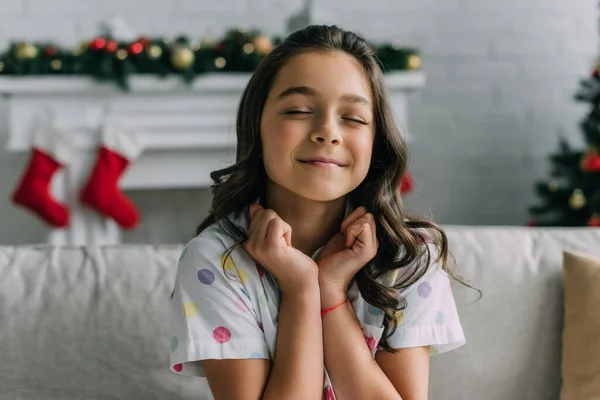 Удовлетворенный ребенок в пунктирной пижаме сидит на диване во время празднования Рождества дома — стоковое фото