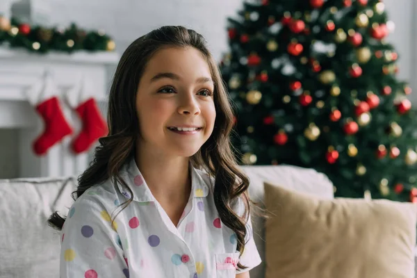 Улыбающийся ребенок в пижаме смотрит в сторону во время празднования Рождества дома — стоковое фото