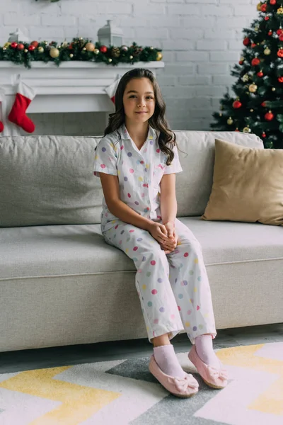 Ragazza sorridente in pigiama punteggiato seduto sul divano vicino al camino sfocato e l'albero di Natale a casa — Foto stock