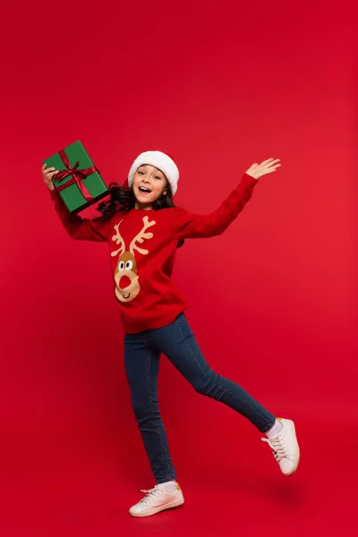 Cheerful girl in Christmas sweater and santa hat holding gift box on red background — Stock Photo