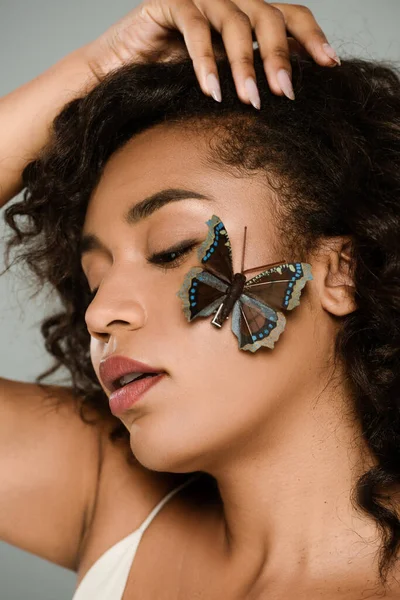 Mujer afroamericana bonita con la mariposa en la mejilla aislada en gris — Stock Photo