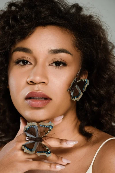 Кучерява афроамериканська жінка з метеликами на руці і обличчям дивиться на камеру ізольовано на сірому — стокове фото