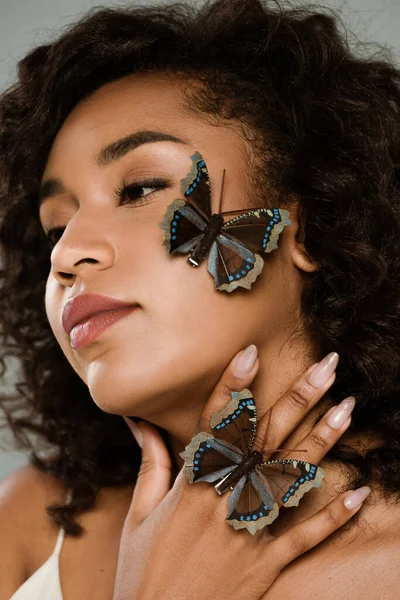 Кучерява афроамериканська жінка з метеликами на руках і обличчі, дивлячись ізольовано на сірому — стокове фото