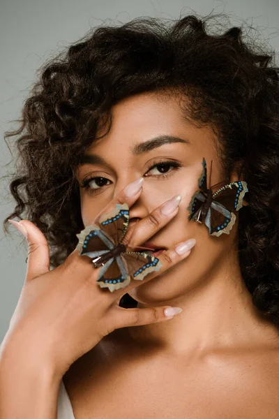 Joven mujer afroamericana con mariposas en la mano y la mejilla cubriendo la cara aislado en gris - foto de stock