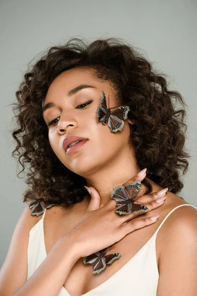 Mujer afroamericana rizada con mariposas en la mano y la mejilla posando aislada en gris - foto de stock