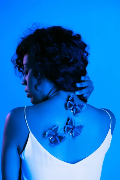 Кучерява афроамериканська жінка з метеликами на спині позує ізольовано на яскраво-синьому — стокове фото
