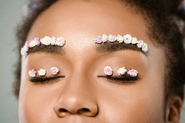 Vista ritagliata di bella donna afroamericana con fiori sulle sopracciglia e occhi chiusi isolati sul grigio — Foto stock
