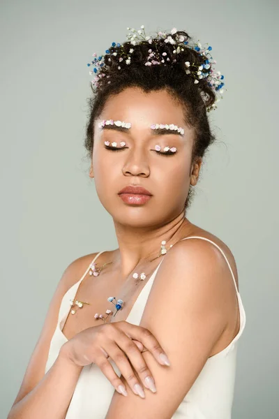 Giovane donna afroamericana con fiori sulle sopracciglia e occhi chiusi in posa isolata sul grigio — Foto stock