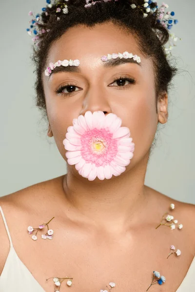 Brunette african american woman with flowers on eyebrows and hair with gerbera in mouth isolated on grey — Stock Photo