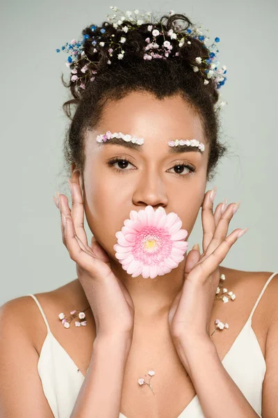 Curly african american woman with flowers on eyebrows and hair with gerbera in mouth isolated on grey — Stock Photo
