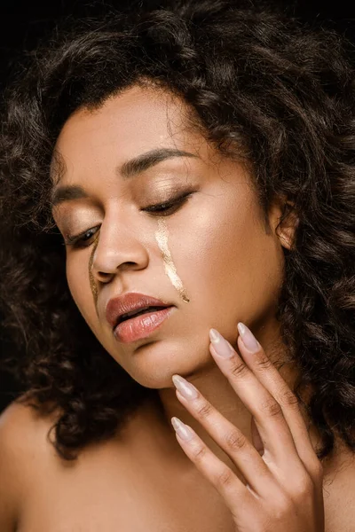 Mulher americana africana com lágrimas douradas nas bochechas tocando rosto isolado no preto — Fotografia de Stock