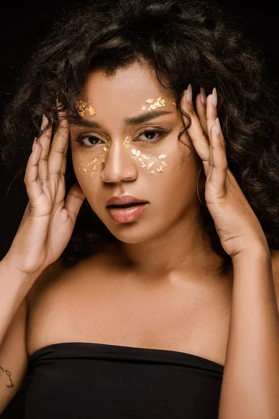 Curly african american woman with golden paint on cheeks looking at camera isolated on black — Stock Photo