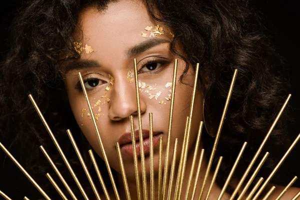 Curly african american woman with golden paint on cheeks looking at camera through crown isolated on black — Stock Photo