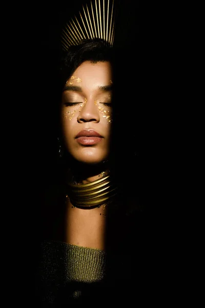 Mujer joven afroamericana con accesorios dorados y pintura en la cara aislada en negro — Stock Photo