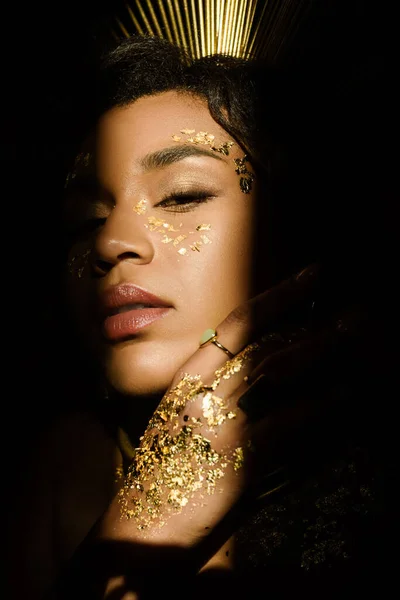 Joven mujer afroamericana con accesorios dorados y pintura en la cara mirando a la cámara a través de la sombra aislada en negro - foto de stock