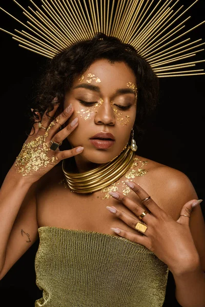 Joven afroamericana mujer con accesorios de oro y pintura en la cara posando aislado en negro - foto de stock