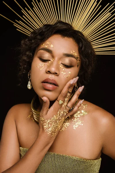 Mujer afroamericana rizada con accesorios dorados y pintura en la cara posando aislado en negro - foto de stock