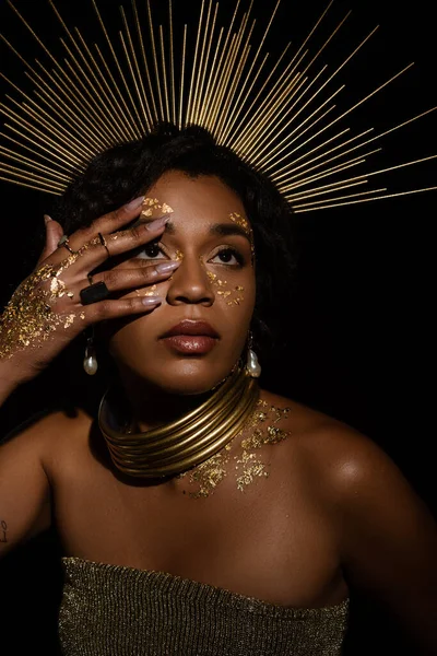 Jeune femme afro-américaine avec des accessoires dorés et de la peinture sur le visage regardant isolé sur noir — Photo de stock