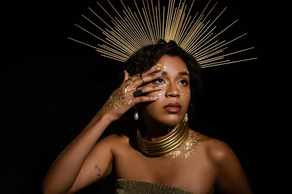 Tatuato donna afroamericana con corona d'oro e vernice sul viso guardando verso l'alto isolato su nero — Foto stock