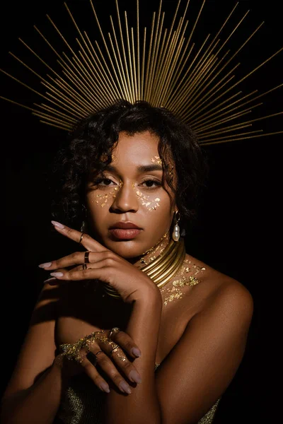 Joven mujer afroamericana con collar de oro y pintura en las mejillas mirando a la cámara aislada en negro - foto de stock