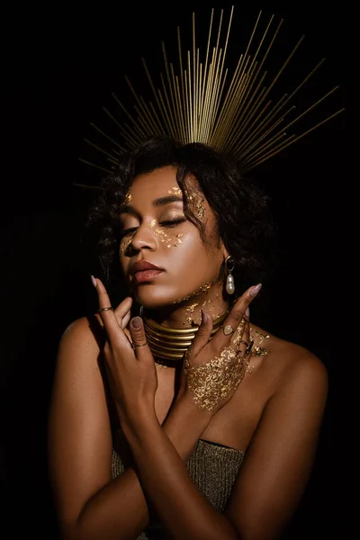 Mujer afroamericana confiada con collar de oro y pintura en las mejillas posando aislado en negro - foto de stock