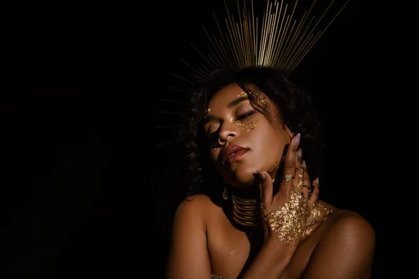 Mujer afroamericana confiada con corona dorada y pintura en la cara posando aislada en negro - foto de stock