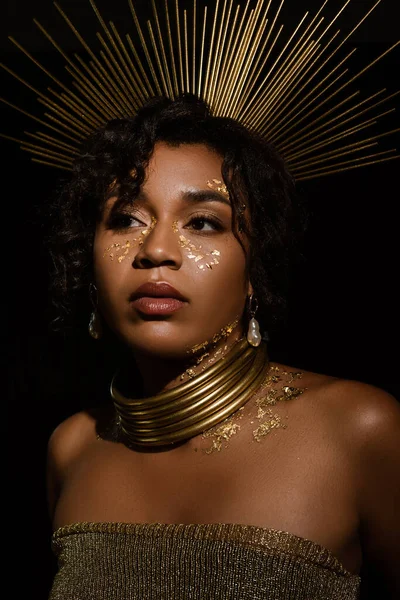 Mujer afroamericana confiada con corona dorada y pintura en las mejillas mirando hacia otro lado aislado en negro - foto de stock