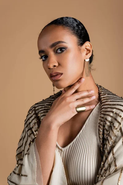 Young african american woman in golden jewelry looking at camera isolated on beige — Stock Photo
