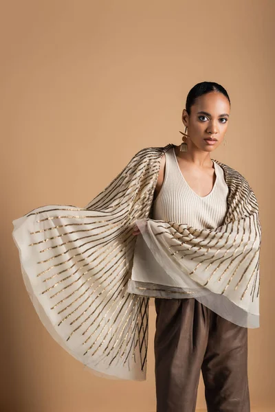 Stylish african american woman in golden jewelry looking at camera and posing in shawl isolated on beige — Stock Photo