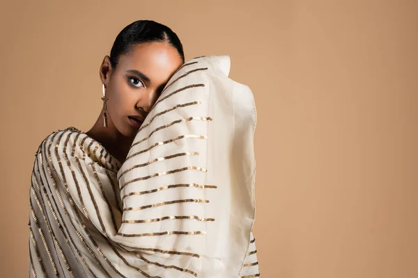 Elegante mujer afroamericana en pendiente de oro mirando a la cámara mientras posando aislado en beige - foto de stock