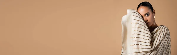 Элегантная африканская американка в золотой сережке смотрит в камеру, позируя изолированно на бежевом, баннере — стоковое фото