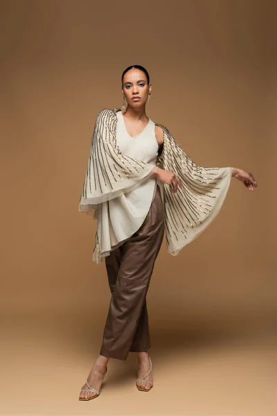 Longitud completa de la elegante mujer afroamericana en chal de oro y pantalones de cuero posando en beige - foto de stock